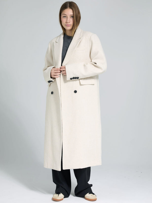 A model showcases a stylish cream-colored long coat with a relaxed fit, featuring black buttons and pockets, by MEOTINE.