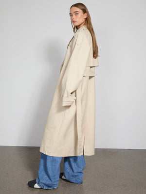 A model showcases a stylish beige trench coat by MEOTINE, paired with wide-leg jeans, set against a minimalist backdrop.