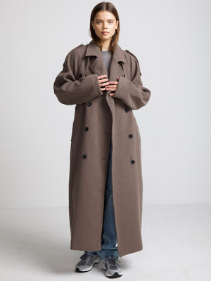 A model wears a long, oversized brown coat with a double-breasted design, styled with casual jeans and sneakers by MEOTINE.