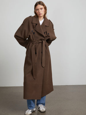 A model wears a stylish brown trench coat with a belt, featuring oversized sleeves, paired with casual jeans and sneakers. MEOTINE.