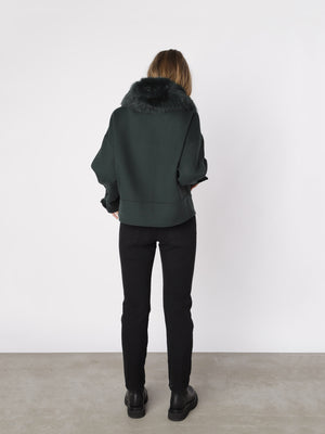 A woman stands with her back to the camera, wearing a dark green MEOTINE jacket with a fur collar, paired with black jeans.