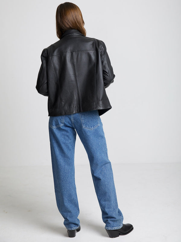 A woman in a black leather jacket and blue jeans stands with her back to the camera, showcasing the stylish MEOTINE outerwear.