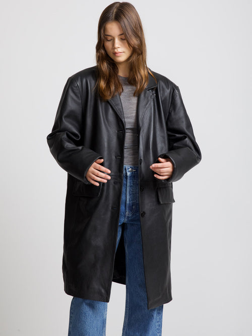 A model wears a stylish black leather coat from MEOTINE, paired with a gray top and blue jeans, showcasing a chic look.