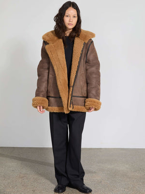 A model wears a cozy brown shearling jacket with a plush collar and cuffs, paired with black wide-leg trousers. MEOTINE style.